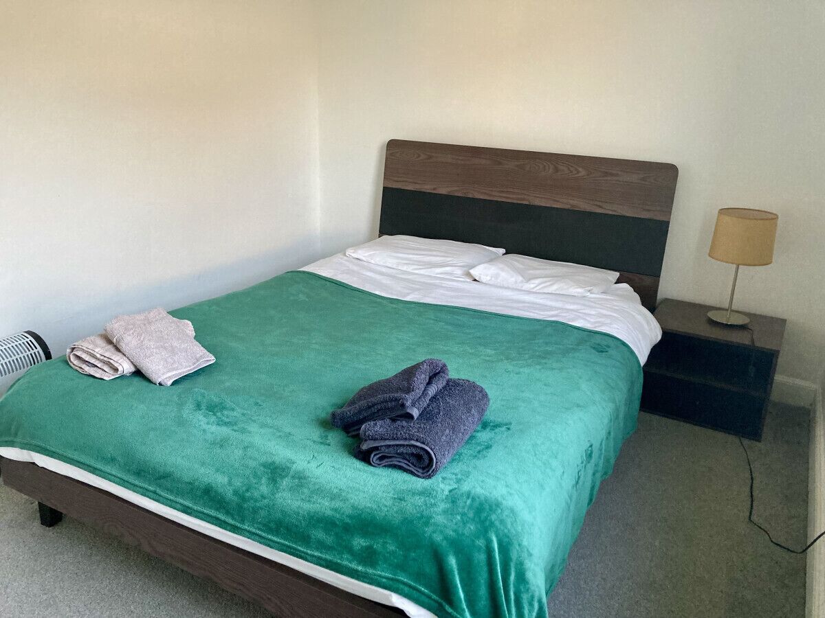 Een net opgemaakt bed met een groene spreidek en twee opgevouwen handdoeken, een bedlamp en een klein nachtkastje.