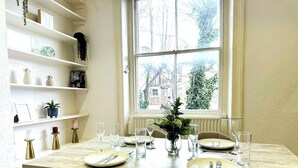 A dining area with a table set for four, a window with a view of trees, and shelves with decorative items. - 2-Bed Maida Vale flat | Little Venice, Paddington (London)