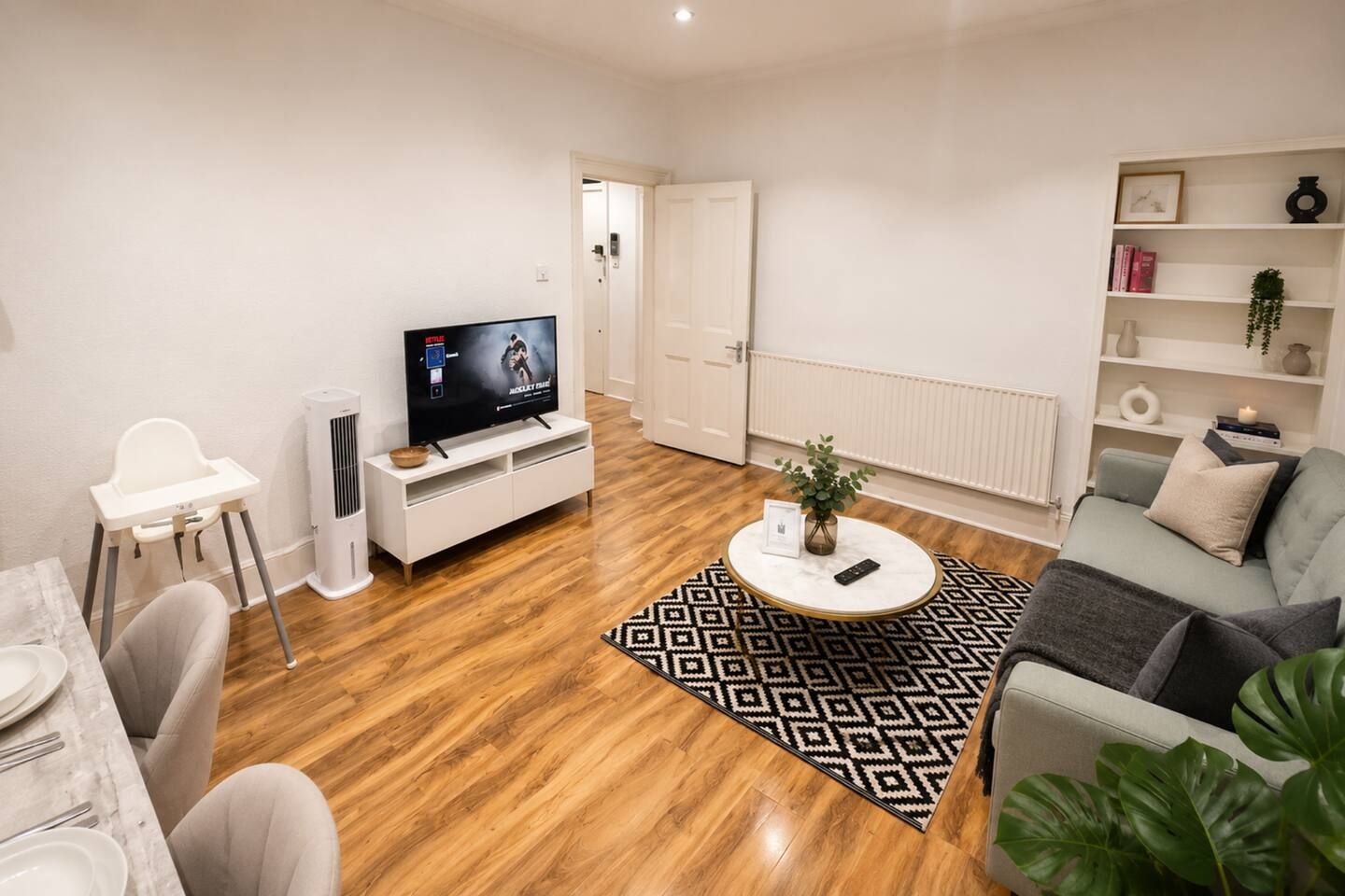 Uma sala de estar com televisão, um sofá branco, uma mesa de centro e um espaço de jantar com cadeiras e uma mesa.
