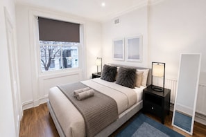 A bedroom with a bed, a nightstand, a lamp, a window with a blind, and a mirror. - 2-bed Maida Vale Flat | little venice, paddington (London)