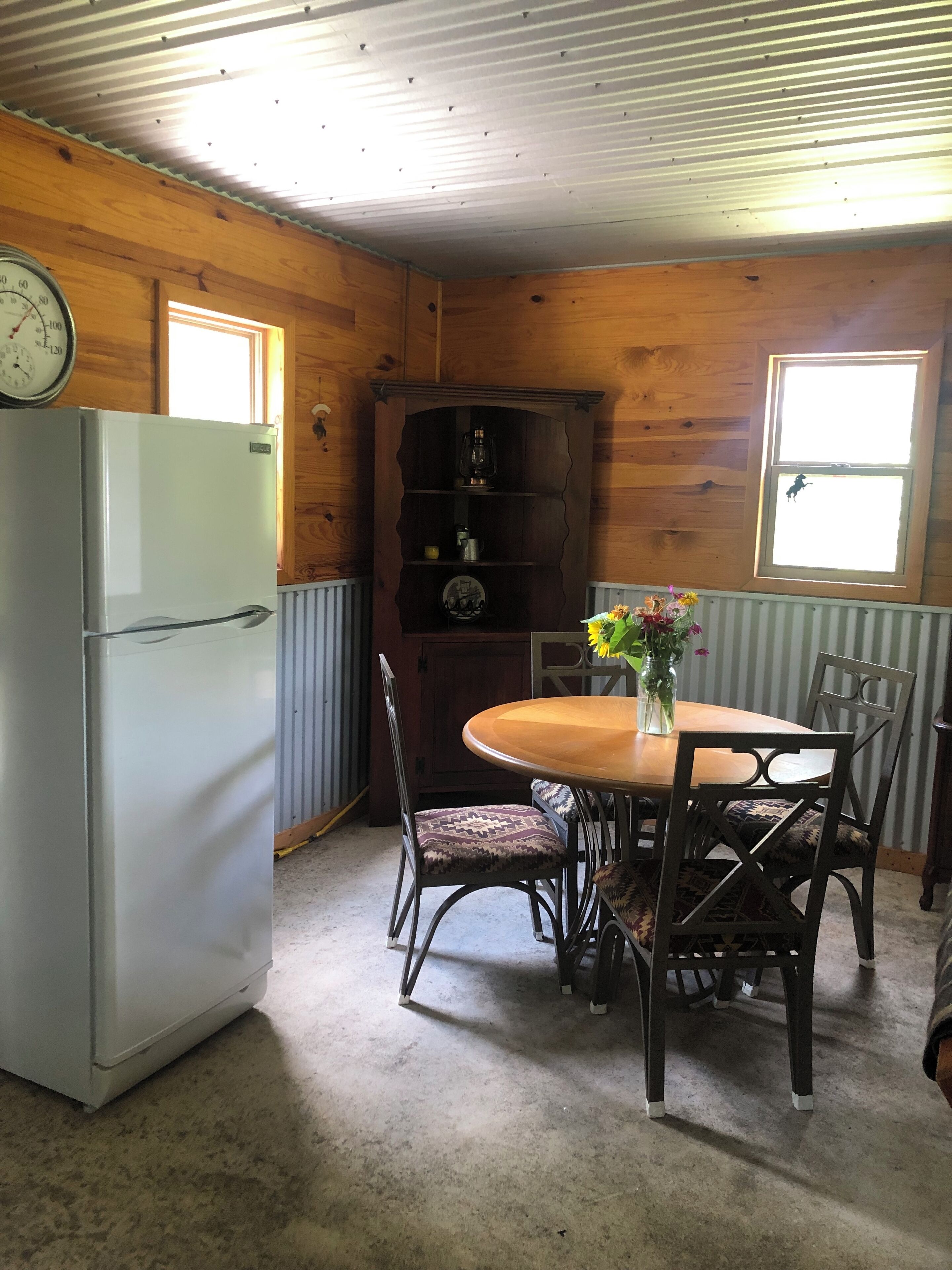 A compact kitchen with a refrigerator, wooden cabinets, a clock, a wooden table with chairs, and a window with a view.