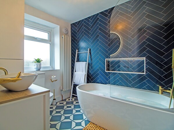 A bathroom with a freestanding bathtub, a white sink with a gold faucet, a window with a plant, and a blue and white patterned floor.