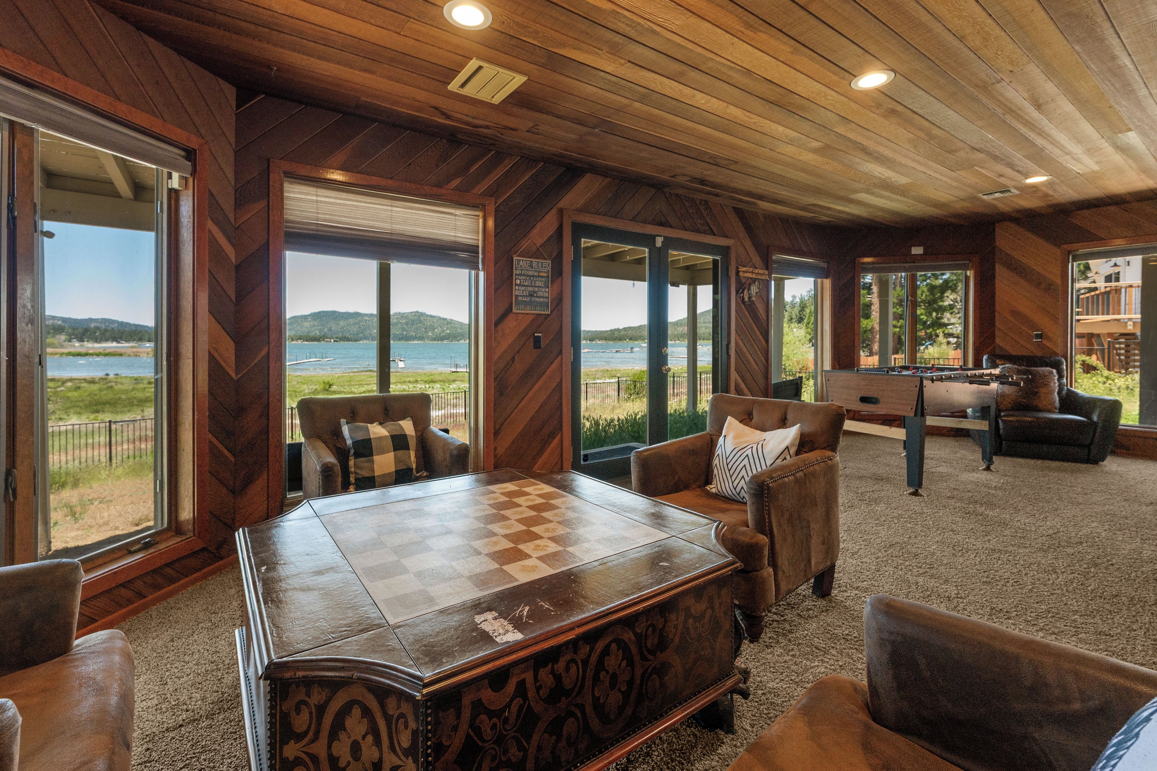 Ein geräumiges Zimmer mit einem großen Holztisch, bequemen Sitzmöglichkeiten und großen Fenstern mit Blick auf einen See und Hügel.