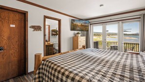 A bedroom with a large bed, a wooden door, a dresser, a mirror, and a view of a lake through large windows.