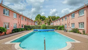 A rectangular swimming pool with a blue cover, surrounded by a brick walkway, and adjacent to pink buildings with multiple windows. - Garden Apartment - UK44654 (Paignton)