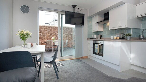 A modern kitchen with white cabinets, a flat-screen TV, and a stone wall visible through the sliding glass door. - Garden Apartment - UK44654 (Paignton)
