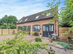 Une maison en briques avec un toit en tuiles, une porte d’entrée et un petit patio aménagé avec du mobilier d’extérieur.