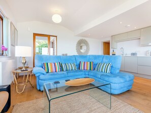 Ein modernes Wohnzimmer mit einem blauen Ecksofa, einem Glastisch und einer Küchenzeile im Hintergrund.