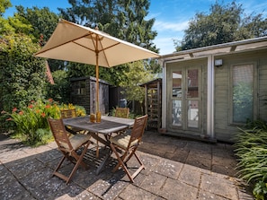 An outdoor patio with a wooden table and chairs, a beige umbrella, and a small wooden shed in the background.