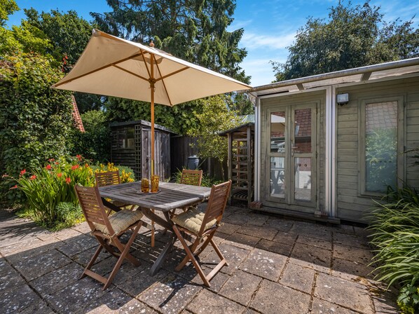 An outdoor patio with a wooden table and chairs, a beige umbrella, and a small wooden shed in the background.