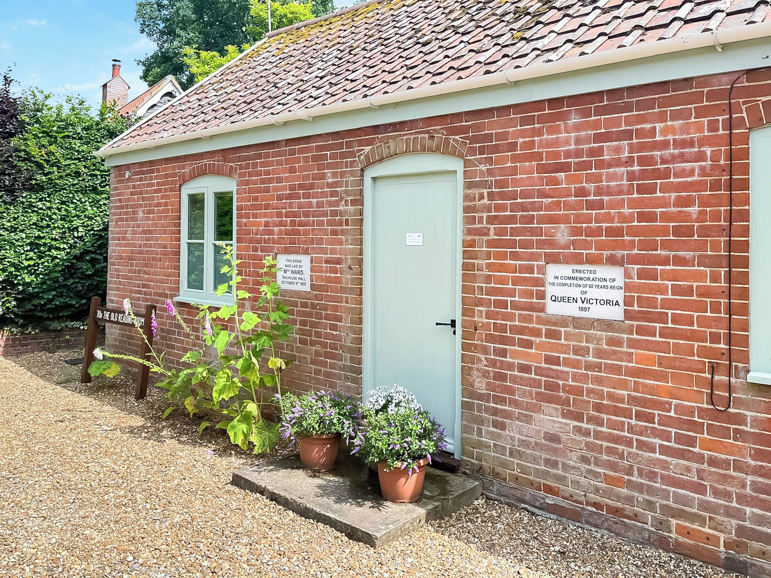 A brick building with a green door, two windows, and potted plants. There are signs on the wall commemorating Queen Victoria's 60th anniversary.