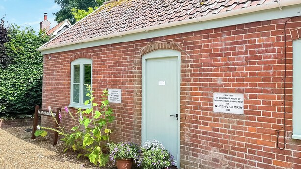A brick building with a green door, two windows, and potted plants. There are signs on the wall commemorating Queen Victoria's 60th anniversary. - The Old Reading Room (Salhouse, near Norwich)