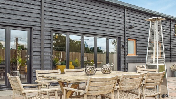 A patio with a wooden table and chairs, a metal chair, and a large window. - 5 bedroom accommodation in Diss (Diss)