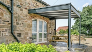 A stone building with a covered patio area, a wooden table, and lush greenery in the foreground. - Discover Yorkshire’s charms from this cosy holiday let. (Warmfield, near Wakefield)