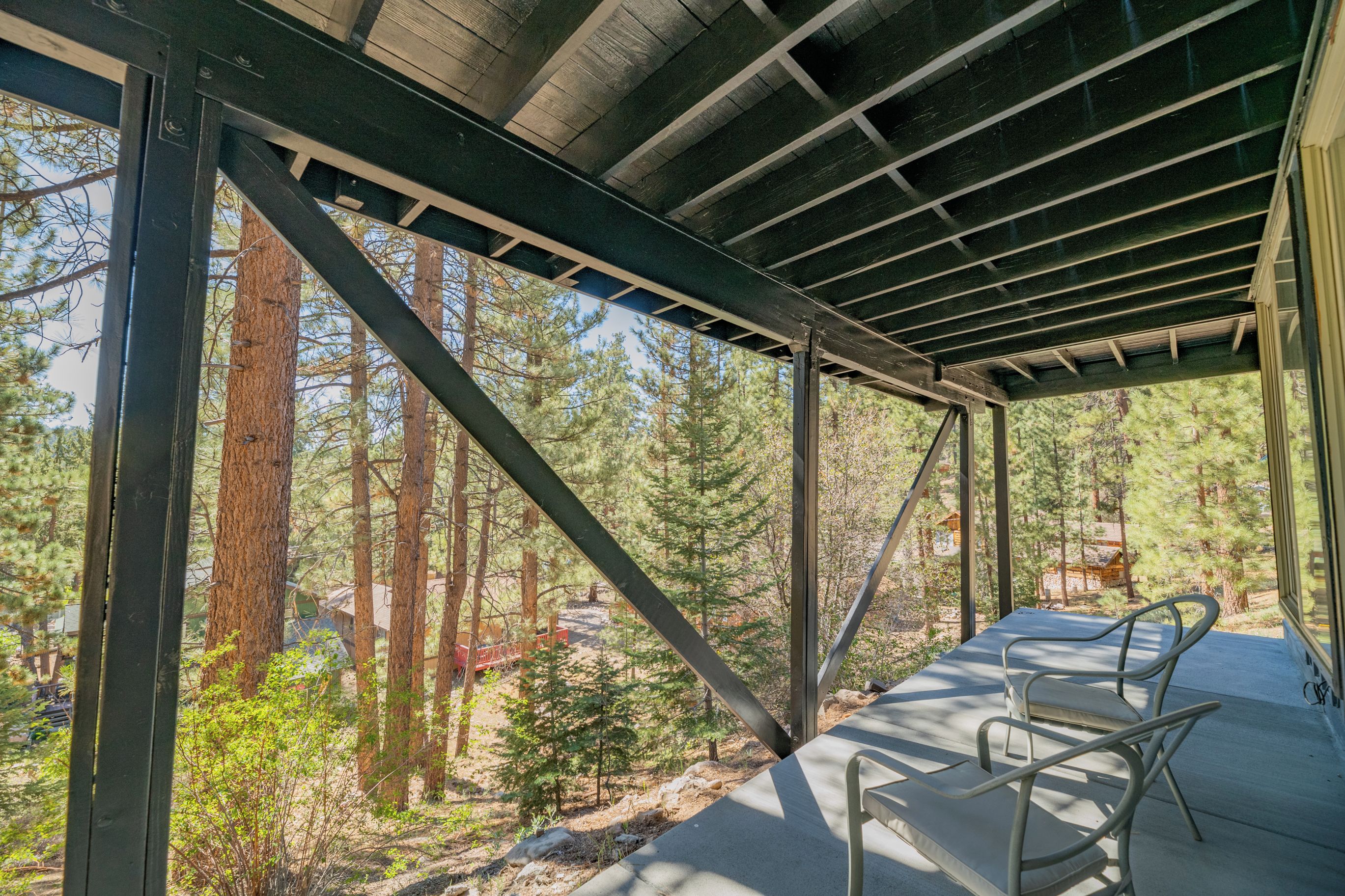 A covered outdoor area with metal beams and a view of a forest. There are two metal chairs on the floor.