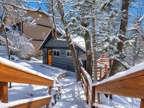 A snow-covered wooden deck leading to a building with a dark roof and a bright orange door. - Forest Escape  (Fawnskin)