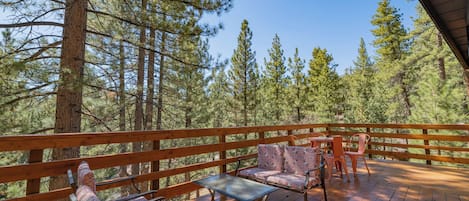 A wooden deck with a table and chairs, surrounded by tall pine trees.