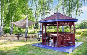 Un kiosque en bois avec un toit bleu et une table de pique-nique, situé dans un espace verdoyant bordé d’arbres.