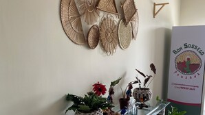 A reception area with a glass table displaying various decorative items, a wall-mounted woven art piece, and a signboard with contact information. - BOM SOSSEGO POUSADA (Delmiro Gouveia)