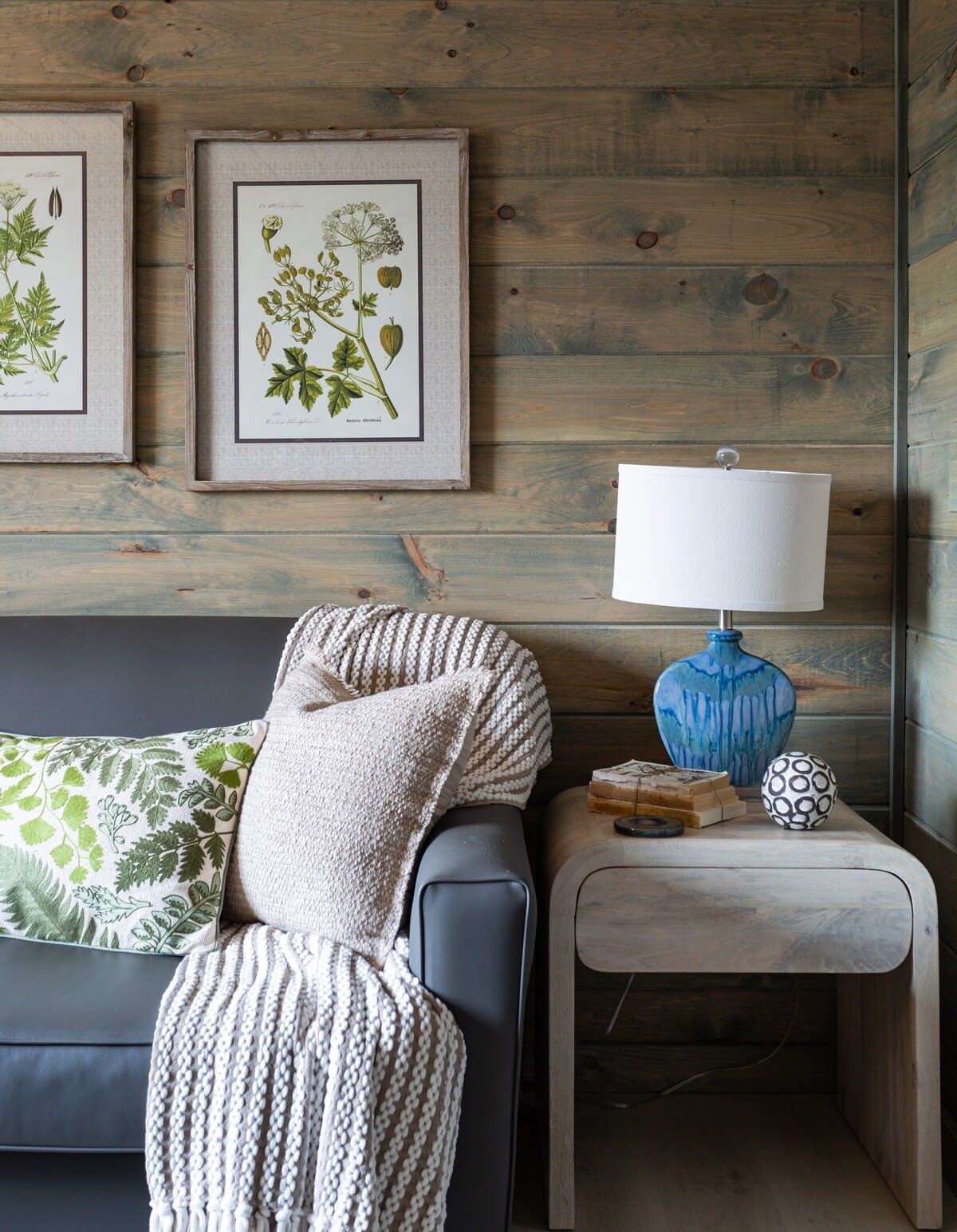 A cozy room with a wooden wall, a black sofa, a patterned throw, a blue lamp, and botanical prints.