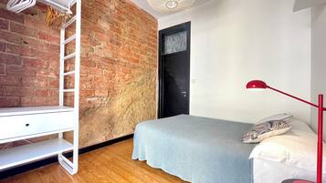 A bedroom with a brick wall, a white wardrobe, a bed with a grey blanket, and a red floor lamp.