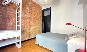 Une chambre avec un mur en briques, une armoire blanche, un lit recouvert d’une couverture grise et une lampe de sol rouge.