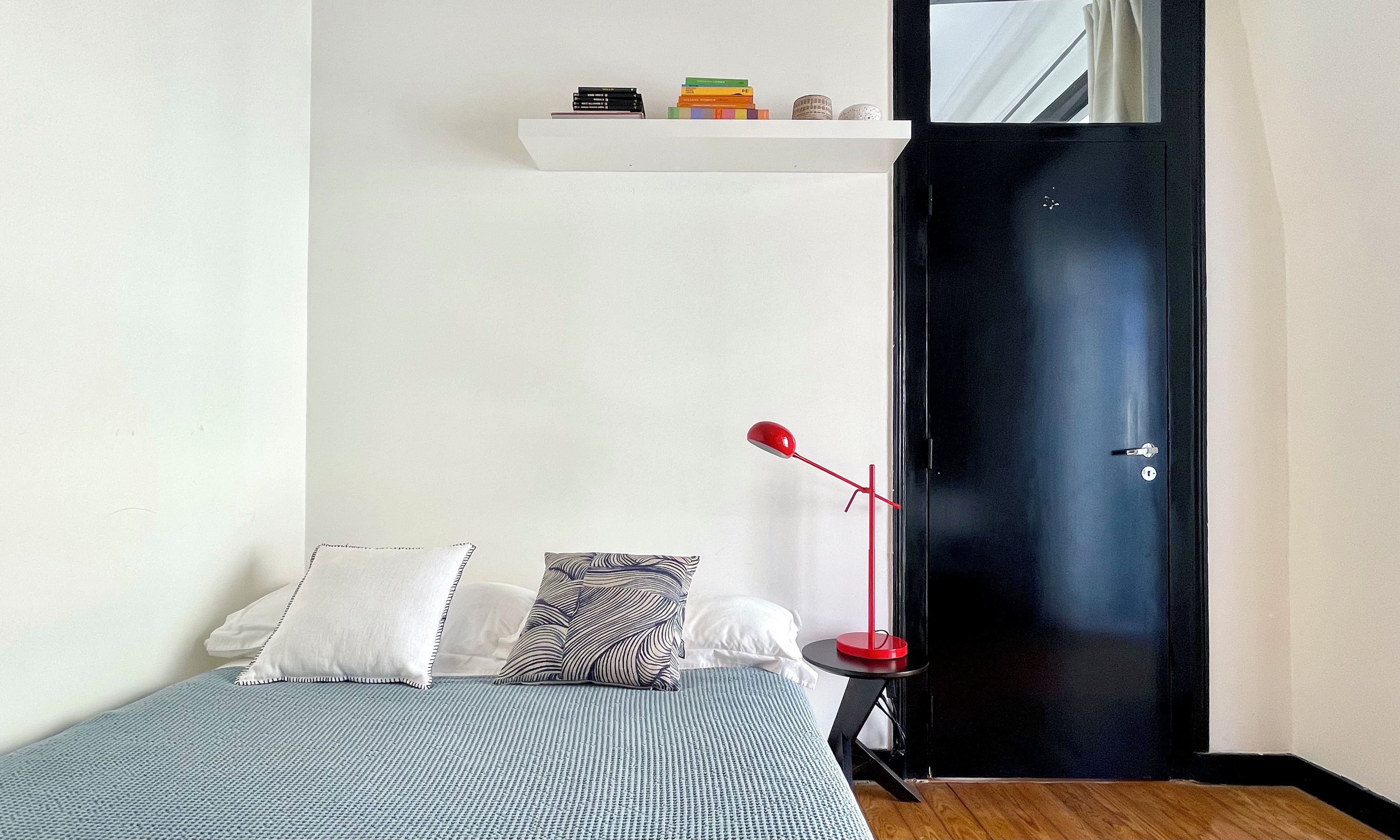 A bedroom with a bed, pillows, a bedside table, a red floor lamp, a black door, and a white shelf with books.