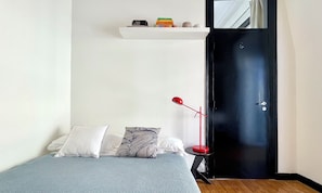 A bedroom with a bed, pillows, a bedside table, a red floor lamp, a black door, and a white shelf with books.
