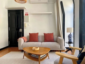 A living room with a sofa, a coffee table, a lamp, and a door marked 'EMERGENCIA 112'.