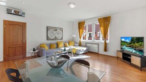 A modern living room with a glass dining table, a sofa with yellow cushions, a television, and a window with curtains. - Exclusive Apartment in Centre (Bratislava)