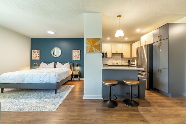 A modern bedroom with a large bed, a small kitchenette, and a dining area with stools.