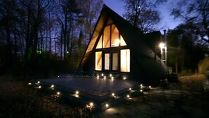 A cabin with a triangular roof, surrounded by trees, and a platform with lights in front.