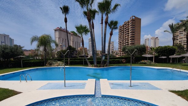 A swimming pool with a clear blue center, surrounded by palm trees and modern buildings in the background. - DOS CALAS sunny apartment (Benidorm)