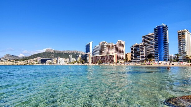 A coastal cityscape with clear blue waters, high-rise buildings, and a mountainous backdrop.
