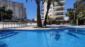 A swimming pool with clear blue water, surrounded by palm trees and a multi-story building in the background.