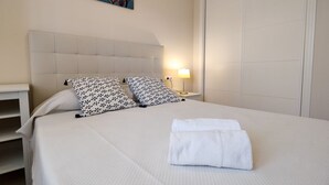 A neatly made bed with a white duvet and two patterned pillows, a bedside lamp, and a wall-mounted artwork.