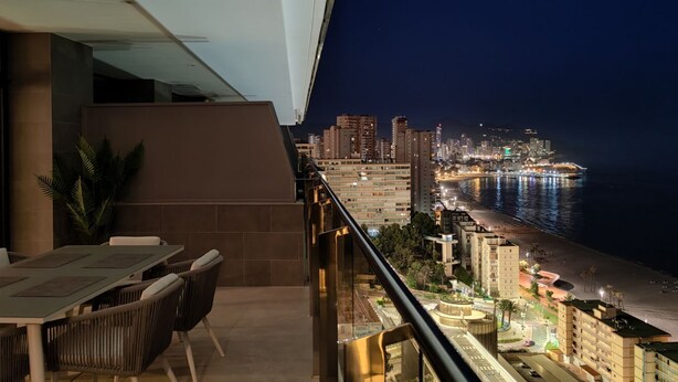 Un balcon doté d’un espace repas offrant une vue sur le paysage urbain et la plage, de nuit.