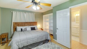 Ein Schlafzimmer mit Bett, Deckenventilator, Nachttisch, einer weißen Tür und einem durch die Tür sichtbaren Badezimmer.