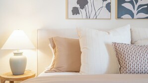 Een netjes opgemaakt bed met een kussenarrangement in wit en beige, een bedlampje en twee ingekaderde bloemkunstwerken aan de muur.