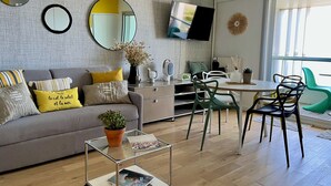 Ein modernes Wohnzimmer mit einem grauen Sofa, gelben Kissen, einem Couchtisch mit Zeitschriften, einem Fernseher an der Wand und einem Essbereich mit Stühlen.