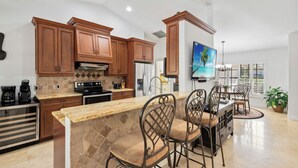 A modern kitchen with wooden cabinets, a granite countertop, and a dining area with metal chairs. - Marco Island Family Retreat  (Marco Island)