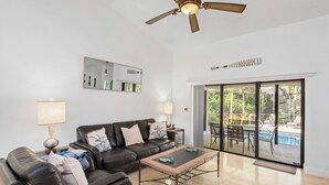 A living room with a black leather sofa, a coffee table, and a ceiling fan. There is a large window with a view of a pool and outdoor furniture. - Marco Island Family Retreat  (Marco Island)