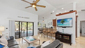 A spacious living room with a ceiling fan, a TV mounted on the wall, a dining area with chairs, and a kitchen area with a TV. - Marco Island Family Retreat  (Marco Island)
