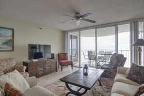 A spacious living room with a flat-screen TV, comfortable seating, a ceiling fan, and a large sliding glass door leading to an outdoor patio with chairs.