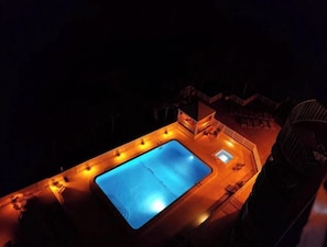 An aerial view of a swimming pool area at night with surrounding deck chairs and a small building.