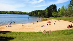 Ein Strand mit einem Volleyballnetz, Menschen, die sich sonnen, und einem See im Hintergrund.