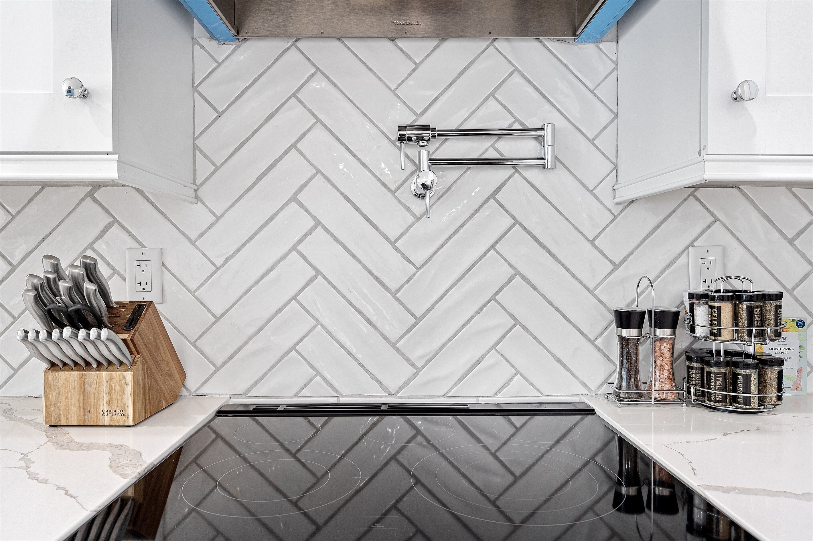A kitchen with a herringbone tile backsplash, a stainless steel faucet, a knife block, and a spice rack.