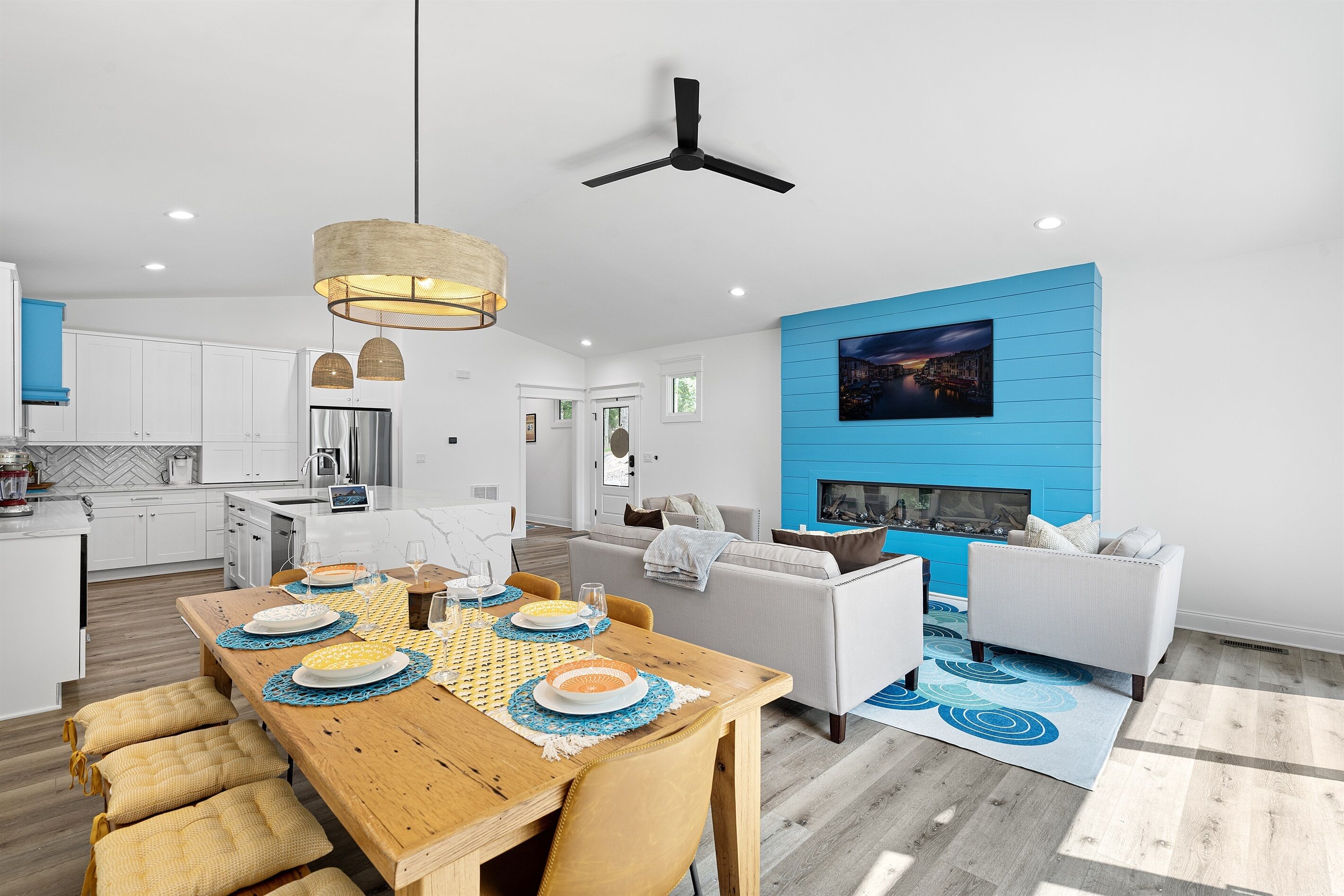 A modern kitchen with white cabinets, a dining area with a wooden table set for four, and a living area with a blue accent wall and a fireplace.