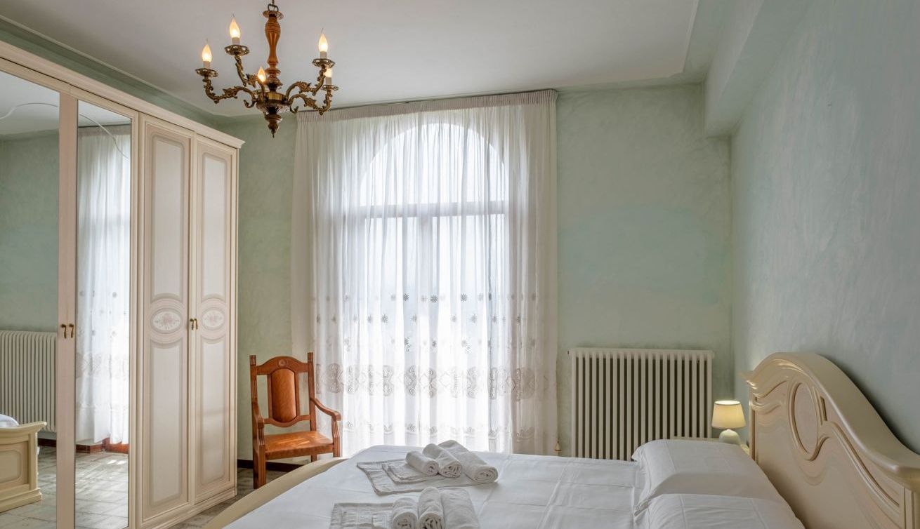 A hotel room with a bed, a chair, a wardrobe, a radiator, and a chandelier.