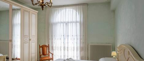 A hotel room with a bed, a chair, a wardrobe, a radiator, and a chandelier.